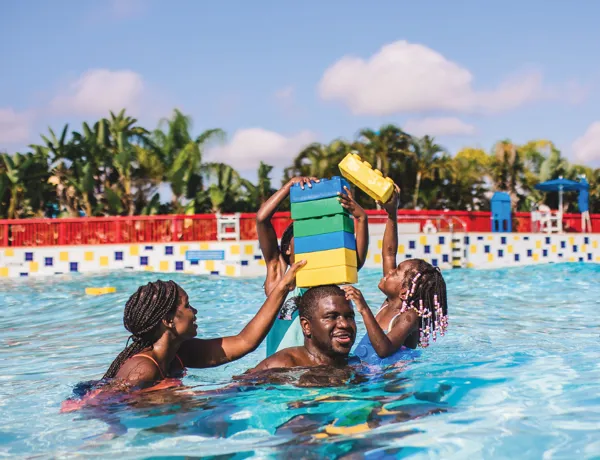 Family of four playing at LEGOLAND Florida Water Park, swimming and stacking oversized LEGO bricks on dad's head.